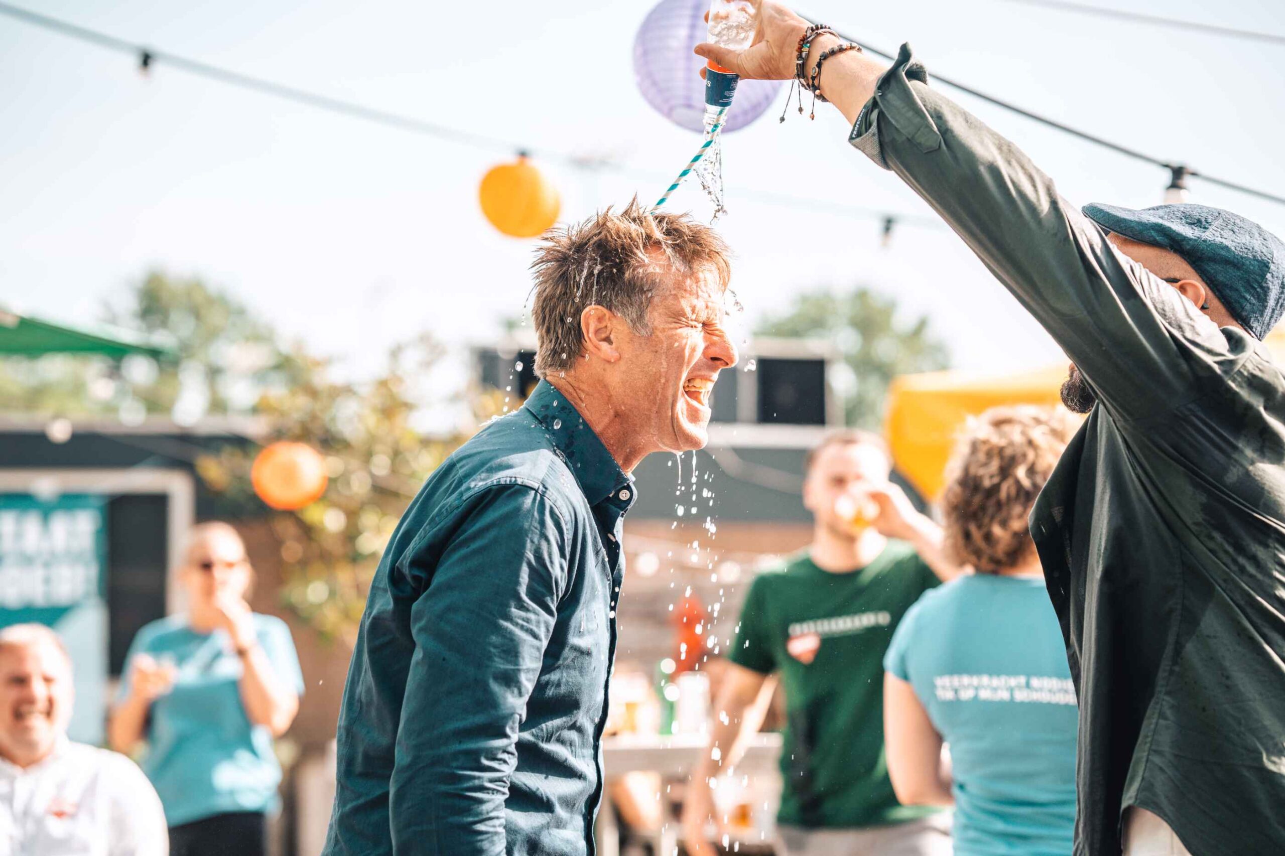 Een man op een winterdipfeestje buiten lacht terwijl water uit een kleurrijke ballon over zijn hoofd wordt gegoten, terwijl de toeschouwers op de achtergrond lachen.