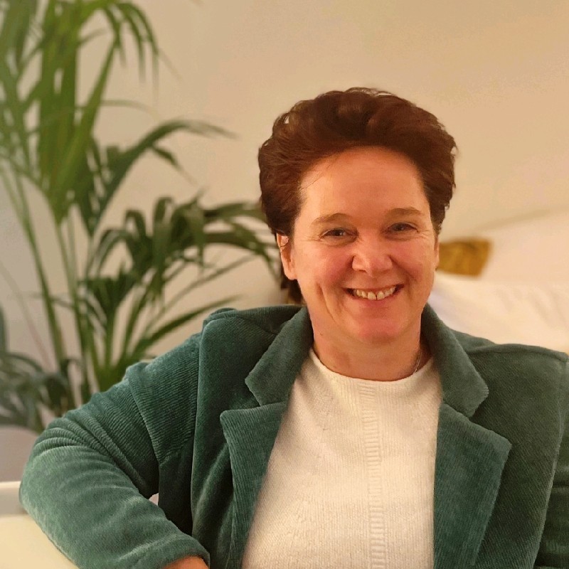 Een vrouw met kort bruin haar glimlacht terwijl ze zit. Ze draagt een groen jasje over een wit topje. Op de achtergrond staat een groene plant, die de serene houding van Marielle Drenth een rustgevende uitstraling geeft.