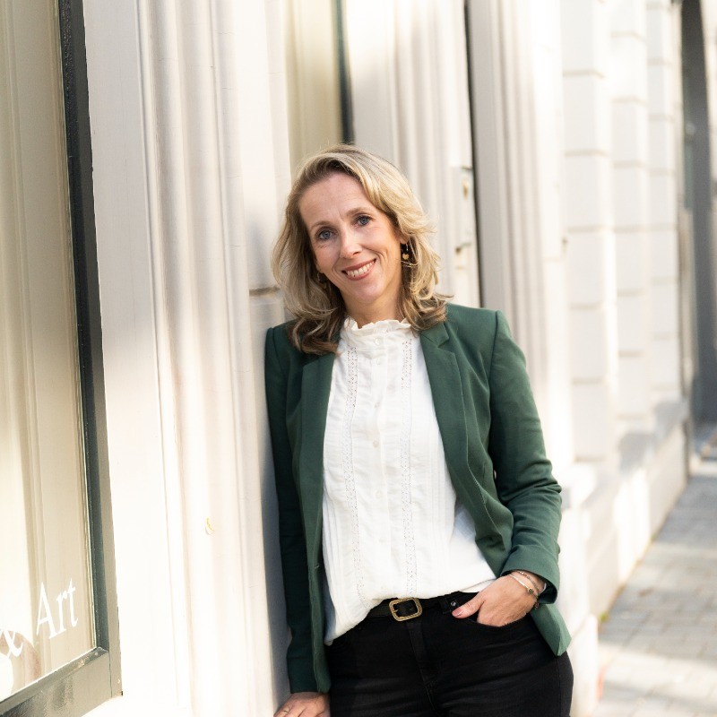 Marion Roebbers, met blond haar op schouderlengte, gekleed in een groene blazer en witte blouse, leunt glimlachend tegen de buitenkant van een gebouw.