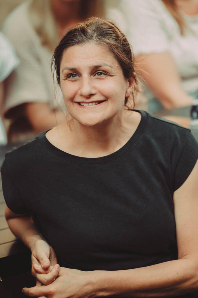 Buiten zit een vrouw met een vriendelijke glimlach, Maaike, in een zwart shirt. Op de achtergrond zijn andere mensen zichtbaar.