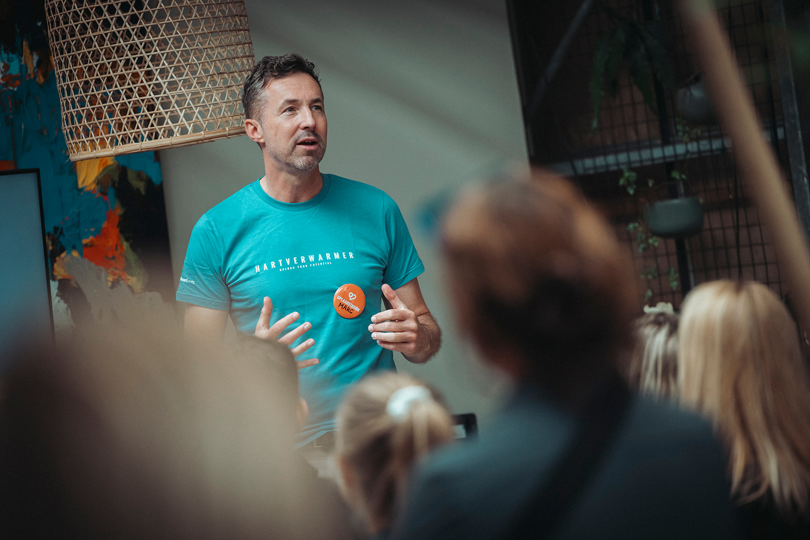Een man in een blauw t-shirt spreekt een publiek toe in een binnenruimte. Op zijn shirt staat een tekst en hij draagt een ronde oranje knoop met de tekst "Wat is duurzame inzetbaarheid." Enkele toeschouwers en een hangende lamp zijn zichtbaar.