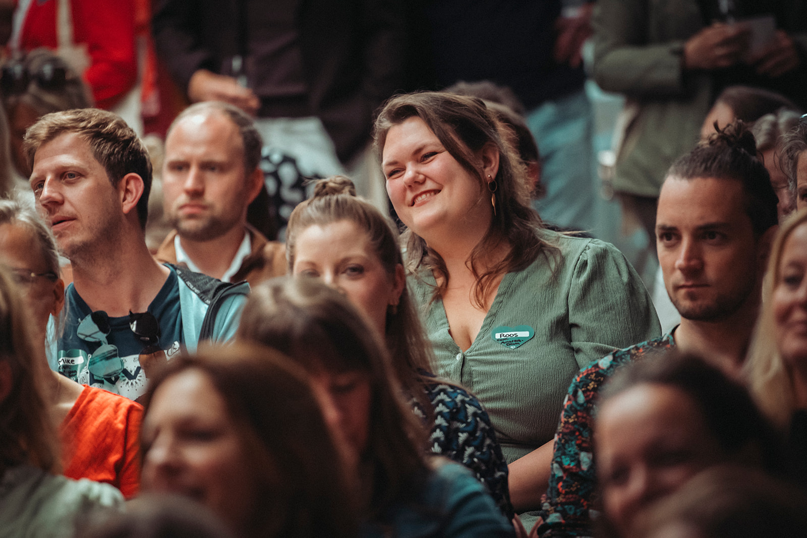 Een groep mensen zit dicht op elkaar in een publiek. Een vrouw in het midden lacht, haar groene outfit weerspiegelt een warme ambiance. Sommige aanwezigen hebben naambordjes.