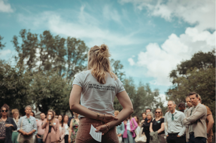 Een vrouw staat met haar rug naar de camera voor een groep buiten, gekleed in een shirt met tekst uit een inspirerend e-book. De lucht is gedeeltelijk bewolkt, wat de thema's vrede en veerkracht weerspiegelt.