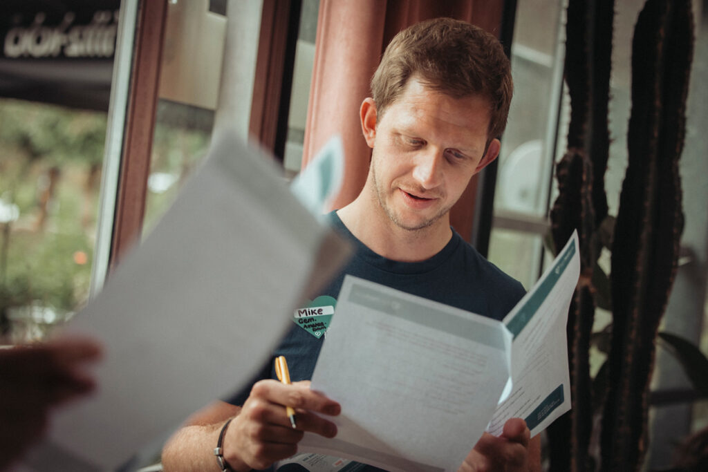 Een persoon die binnenshuis bij een raam met gordijnen staat, houdt papieren en een pen vast en lijkt geconcentreerd een e-book te lezen. Hij straalt een sfeer van veerkracht uit.