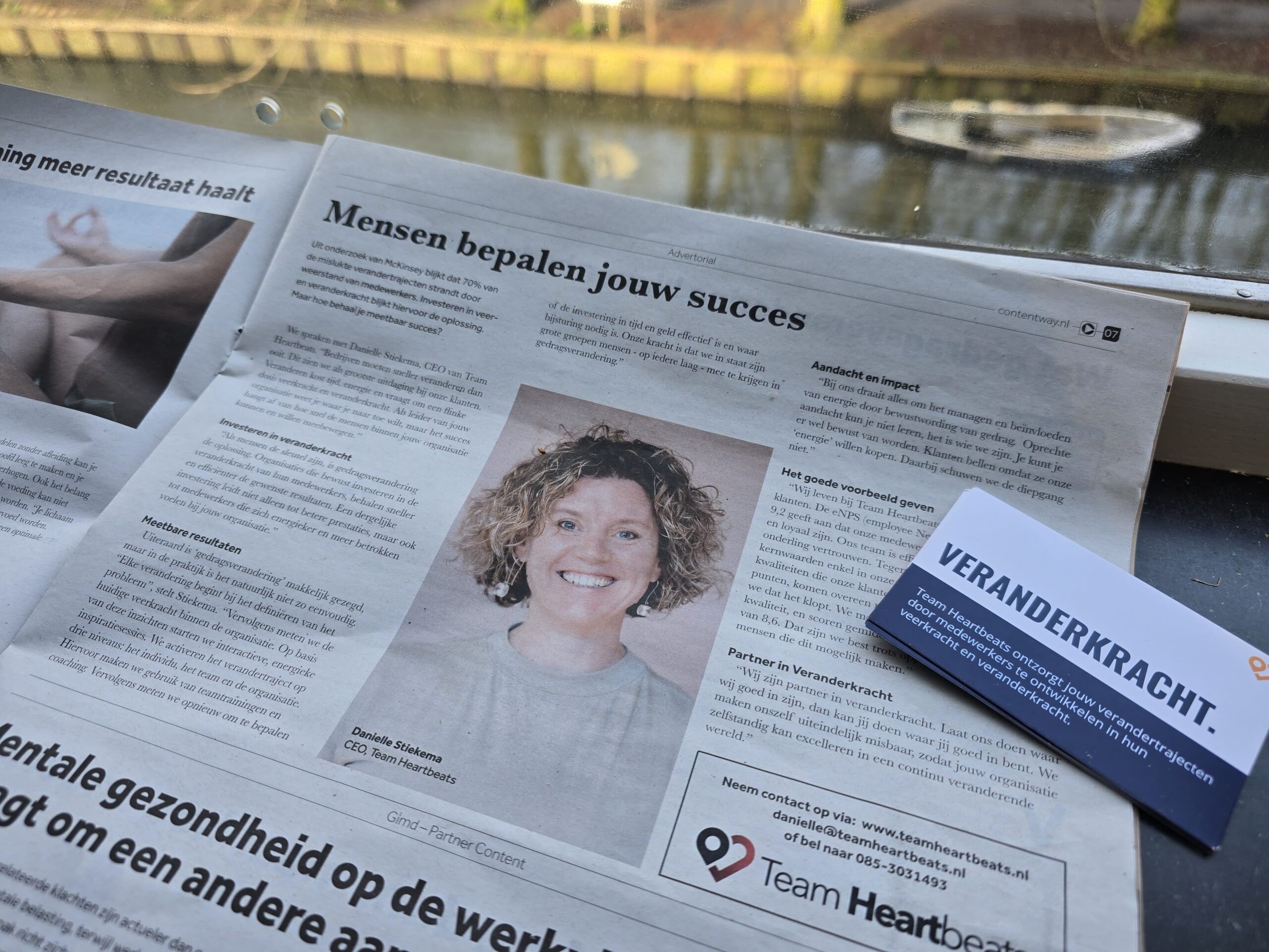 Een krant op een vensterbank toont een artikel getiteld "Mensen bepalen jouw succes", vergezeld door een portret van een lachend persoon. Ernaast benadrukt een visitekaartje met het woord "VERANDERKRACHT" de kracht van transformatie.