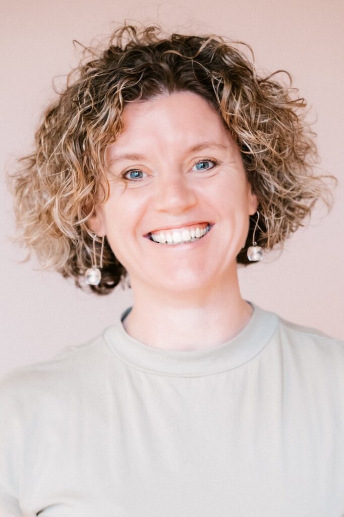 Een glimlachende persoon met krullend haar straalt inspiratiekracht uit terwijl hij een lichtgekleurd shirt en bolvormige oorbellen draagt, poserend tegen een effen achtergrond.