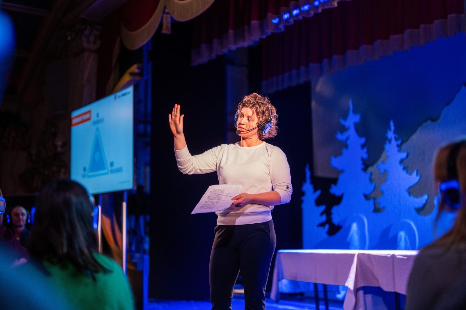Een spreker leidt een sessie met een koptelefoon, draagt een headset en gebaart met papieren op het podium voor een publiek, terwijl een presentatiedia op de achtergrond verschijnt.
