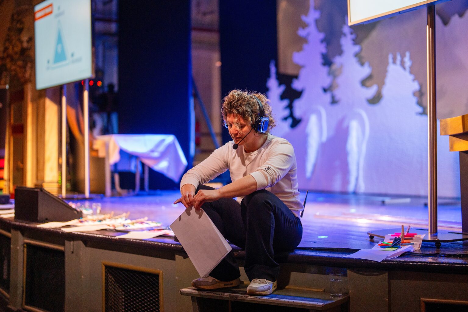 Een persoon geniet van een koptelefoonsessie, zittend op een podiumtrap met papieren in de hand, omringd door presentatieschermen, kunstbenodigdheden en podiumdecoraties met een winters thema op de achtergrond.