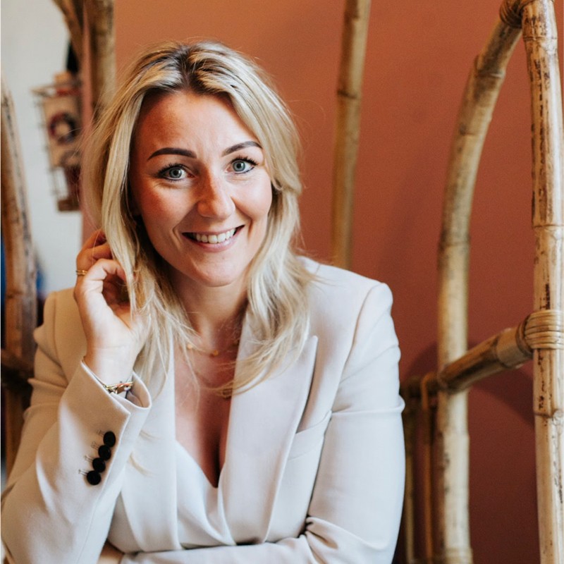 Een vrouw met lang blond haar in een lichtgekleurde blazer glimlacht terwijl ze voor bamboestokken en een rozeachtige muur zit.