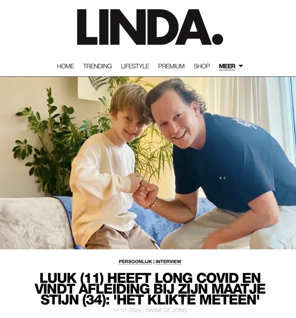 Een jonge jongen en een volwassen man, beiden Handjehelpen vrijwilligers, zitten glimlachend en hand in hand op een bank voor een foto in een lichte woonkamer-genietend van een mooi moment samen.