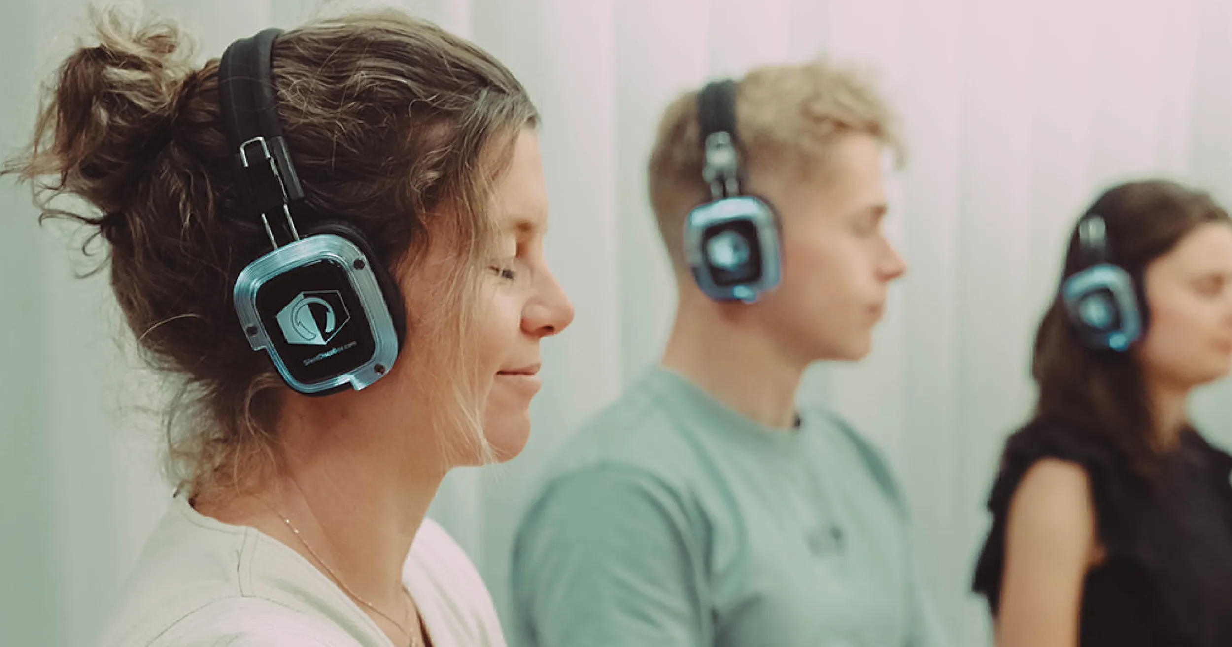 Drie mensen zitten met gesloten ogen op een rij en dragen een grote koptelefoon. Ze lijken in een ontspannen, meditatieve staat van ontspanning te verkeren.